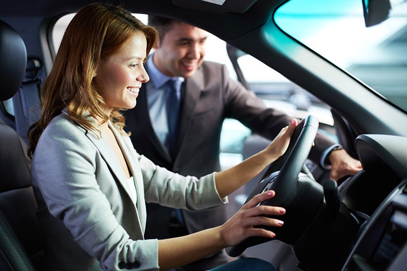 Smiling woman in driver’s seat with cheerful salesman outside